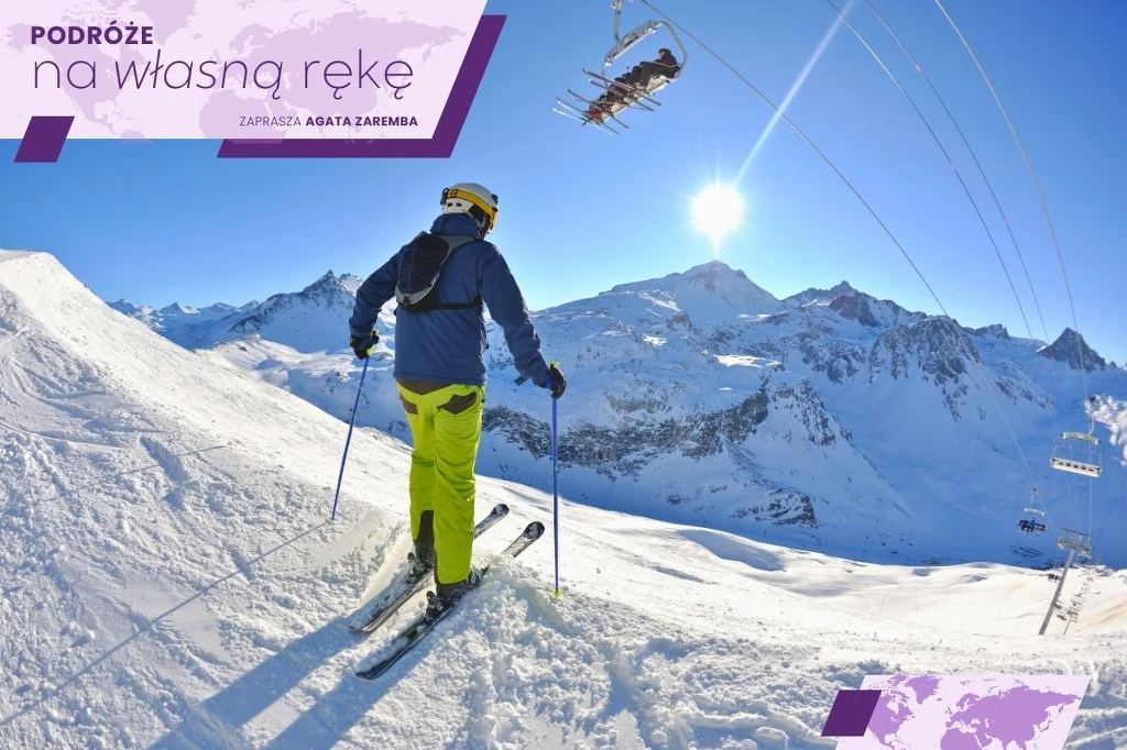 Narciarz ubrany w żółte spodnie i kask stoi na szczycie ośnieżonego stoku, wokół rozciągają się wysokie, górskie szczyty oświetlone słońcem, po prawej stronie widoczny wyciąg krzesełkowy.