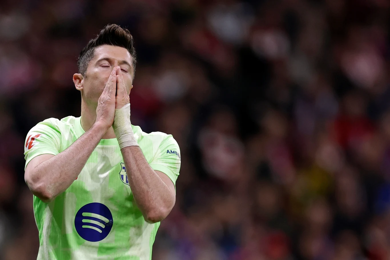 Robert Lewandowski Piłkarz w jasnej koszulce z logo sponsora trzyma dłonie przy twarzy w geście rozczarowania lub żalu, tło rozmyte z widoczną publicznością na trybunach.