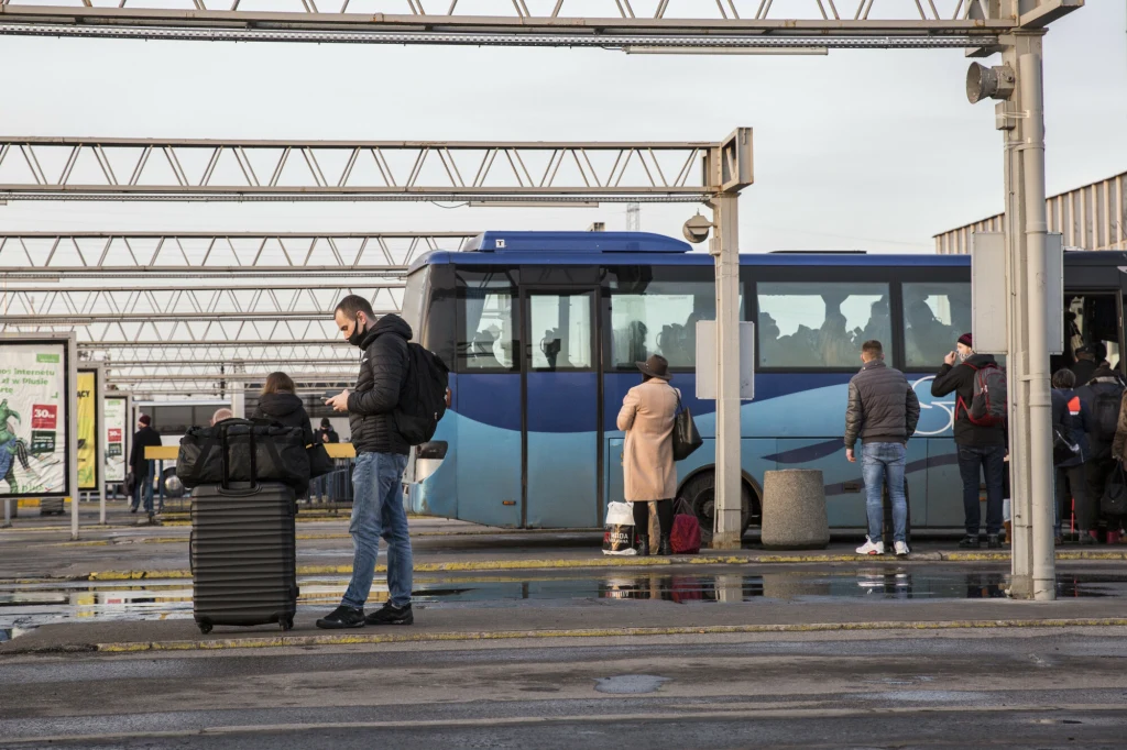 Ludzie oczekujący i wsiadający do niebieskiego autobusu na miejskim dworcu autobusowym, część osób stoi z walizkami i plecakami, na murawie widoczne kałuże po deszczu.