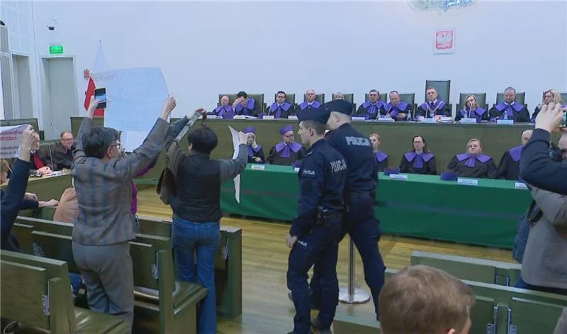 Grupa sędziów siedzących za długim stołem w togach, przed nimi osoby protestujące z podniesionymi transparentami, dwóch policjantów podejmuje interwencję, sala o oficjalnym charakterze z godłem i flagą Polski.