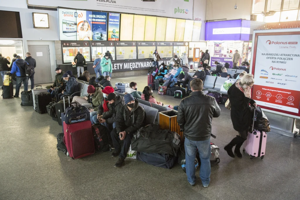 Ludzie siedzący na ławkach i podłodze z walizkami w hali dworca autobusowego, niektórzy wyglądają na zmęczonych lub oczekujących, w tle tablice odjazdów i kasa biletowa.