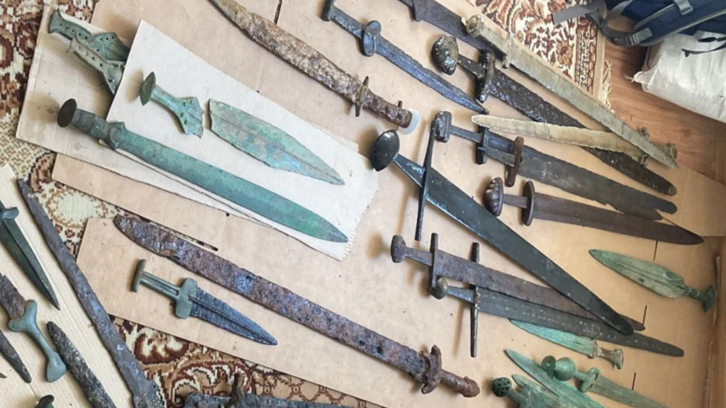 Kolekcja starożytnych mieczy i sztyletów wykonanych z metalu, starannie ułożona na kartonach i dywanie. Różnorodne kształty i stopnie zużycia ostrzy podkreślają ich wiek oraz różnorodność historyczną.