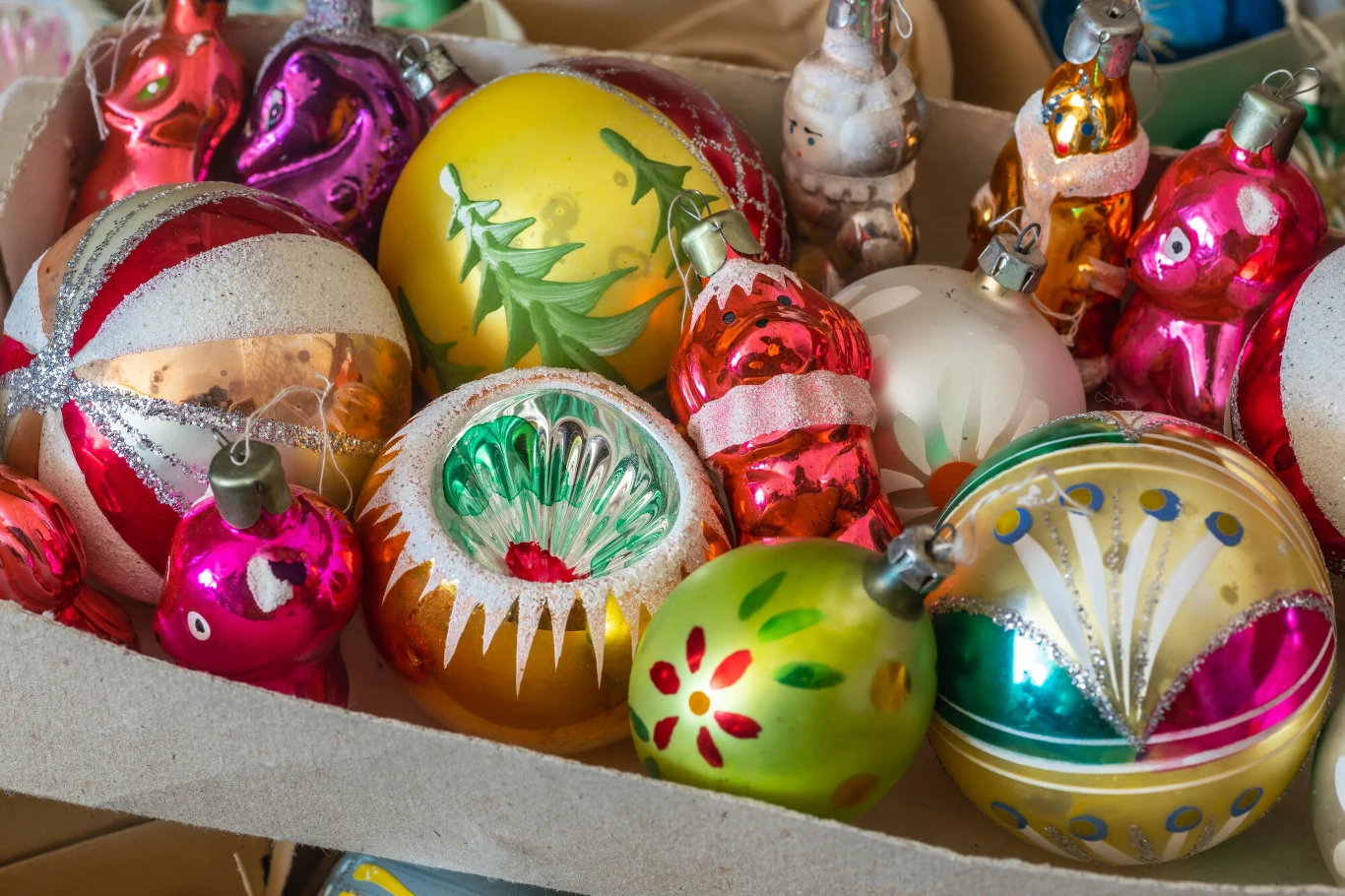 Kolorowe, szklane bombki choinkowe w różnych kształtach i wzorach leżące razem w kartonowym pudełku. Niektóre bombki ozdobione brokatem, inne mają ręcznie malowane motywy świąteczne.