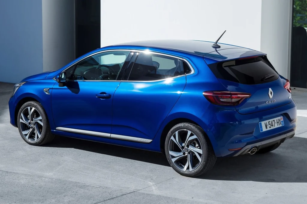 Niebieski samochód marki Renault Clio zaparkowany na jasnej powierzchni przy nowoczesnym budynku, widoczne tylne światła, refleksy na lakierze oraz eleganckie felgi.