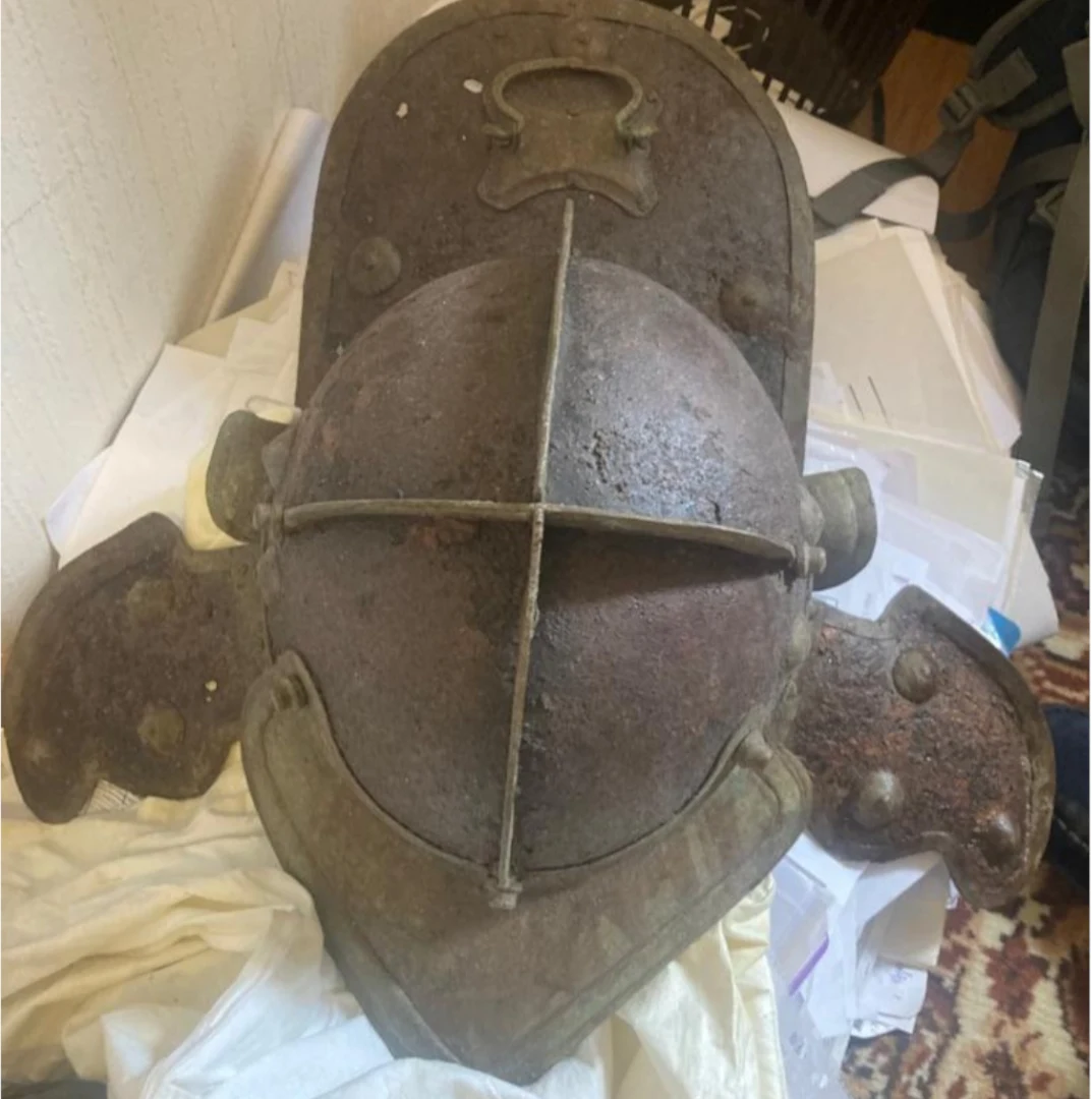 Zardzewiały, średniowieczny hełm rycerski z metalowymi wzmocnieniami i elementami ochronnymi leży na stercie białych i żółtych tkanin oraz dokumentów biurowych.