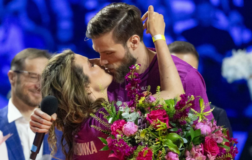 Para w objęciach całuje się na scenie, kobieta trzyma duży bukiet kolorowych kwiatów, wokół obecni są inni ludzie, a w tle widoczne jest rozmazane niebieskie oświetlenie widowiska.