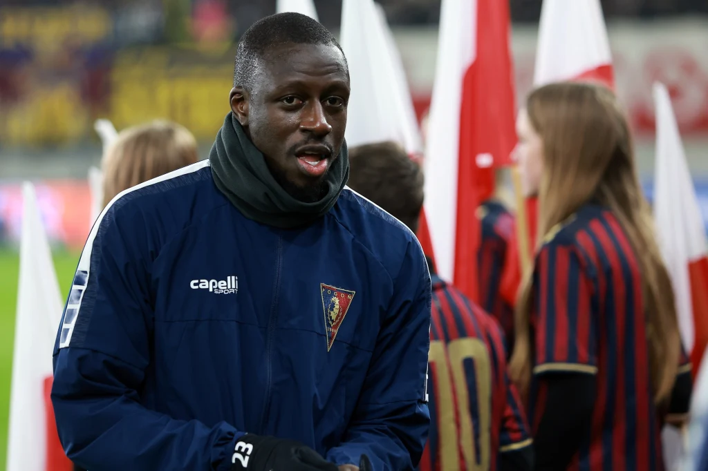 Benjamin Mendy Piłkarz w granatowej kurtce z emblematem klubu sportowego na piersi stoi na tle innych osób trzymających polskie flagi, w tle widać kilka osób w pasiastych strojach.