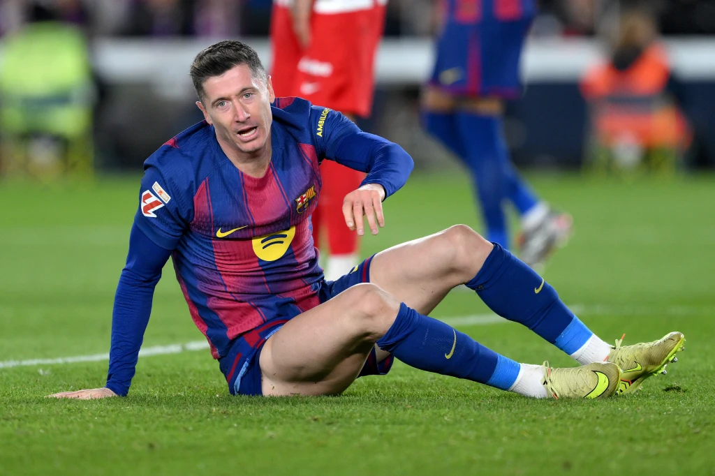 Robert Lewandowski Piłkarz drużyny FC Barcelona siedzi na murawie boiska w stroju meczowym, z widocznym wyrazem zmęczenia lub bólu na twarzy, wokół zamazany tłum oraz fragmenty innych zawodników.