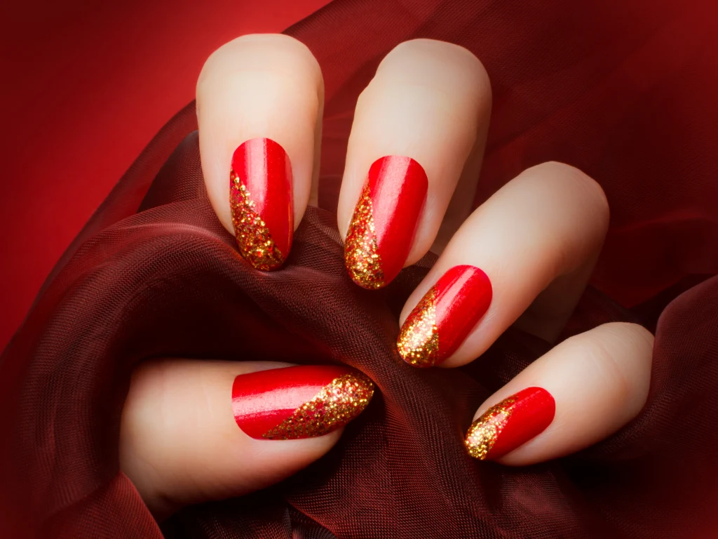 Świąteczne paznokcie, które podkręcą każdą stylizację Dłoń z paznokciami pomalowanymi na czerwono, ozdobionymi złotym brokatowym zdobieniem na końcach, spoczywa na ciemnoczerwonej tkaninie, tworząc efekt elegancji i luksusu.