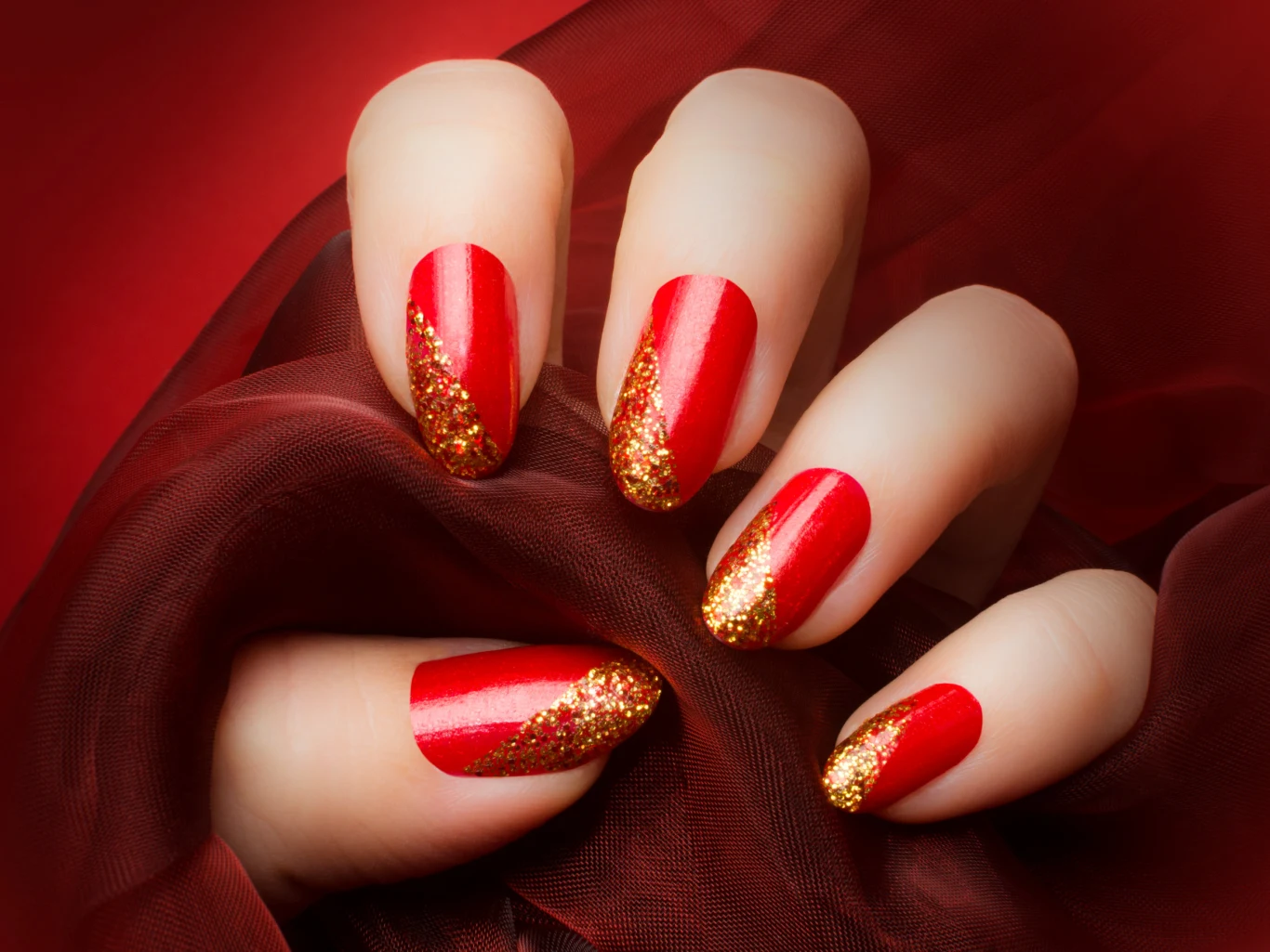 Dłoń z paznokciami pomalowanymi na czerwono, ozdobionymi złotym brokatowym zdobieniem na końcach, spoczywa na ciemnoczerwonej tkaninie, tworząc efekt elegancji i luksusu.