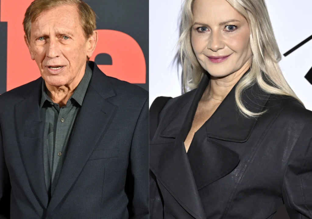 Dwie osoby ubrane elegancko, starszy mężczyzna z krótkimi włosami w garniturze stoi po lewej stronie, kobieta o jasnych, długich włosach i delikatnym makijażu, ubrana w czarną marynarkę, znajduje się po prawej stronie