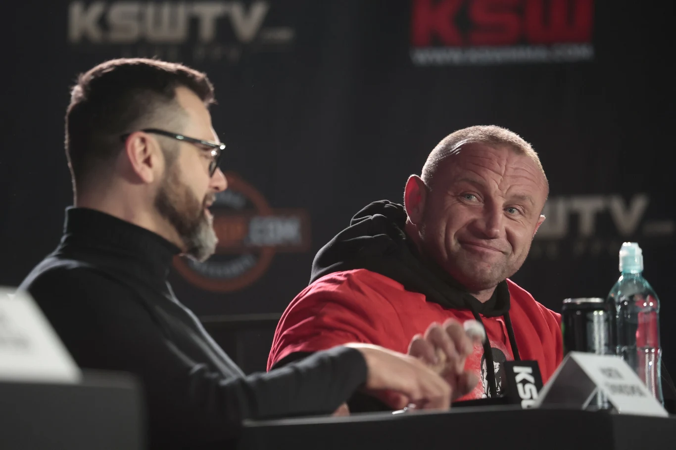 Dwóch mężczyzn siedzi przy stole podczas konferencji prasowej KSW, jeden ubrany w czerwony dres z szerokim uśmiechem na twarzy, obok butelka wody i mikrofon, w tle logo federacji.