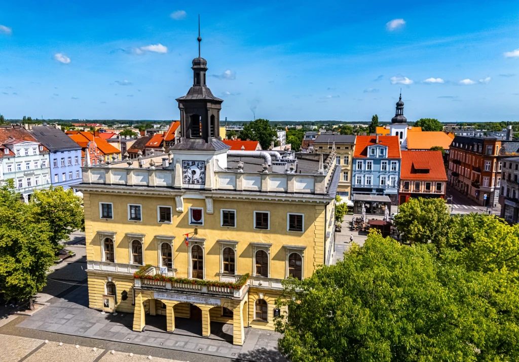 Zabytkowy ratusz z wieżą zegarową w centrum miasta, otoczony kolorowymi kamienicami oraz zielenią drzew, jasne niebo podkreśla malowniczość miejskiego pejzażu.