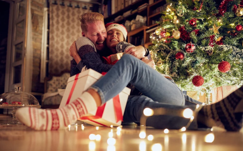 Para w świątecznych swetrach i skarpetkach siedzi przy udekorowanej choince, obejmując się radośnie i trzymając prezent owinięty w biały papier z czerwoną wstążką. Na podłodze wokół nich leżą świąteczne lampki i inne prezenty.