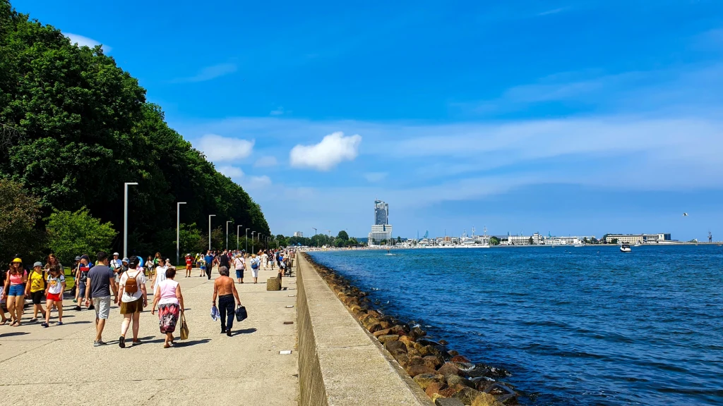 Szeroka promenada nadmorska w słoneczny dzień, po lewej stronie liczne drzewa, po prawej woda oraz daleka panorama miasta z widocznym wysokim budynkiem. Wiele osób spaceruje w letnich ubraniach, ciesząc się pogodą.