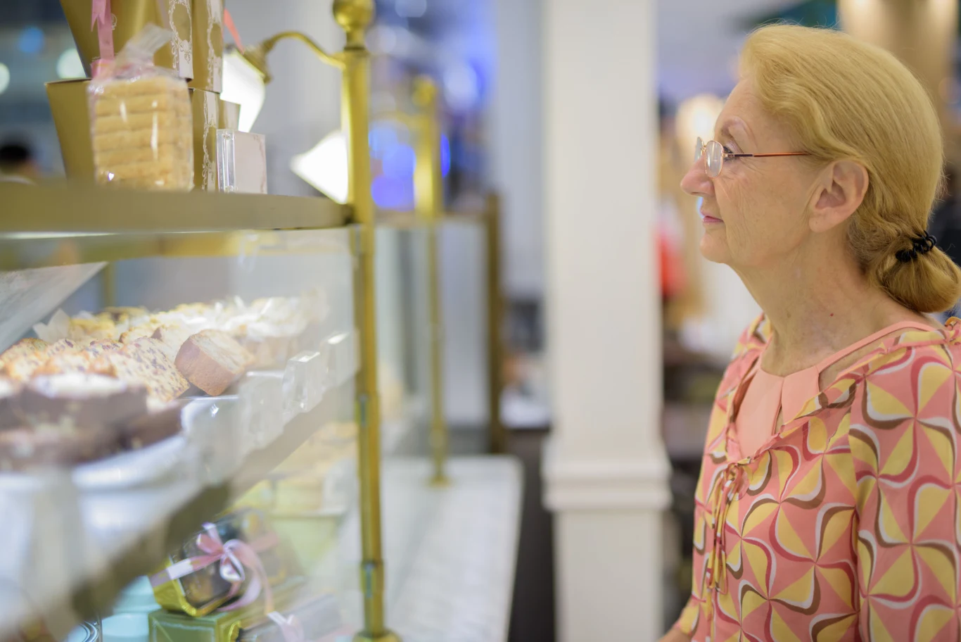 Starsza kobieta uważnie przygląda się ciastom wyłożonym w szklanej gablocie cukierni. W tle rozmyty wystrój wnętrza sklepu, a kobieta nosi okulary oraz ubranie z kolorowym wzorem.