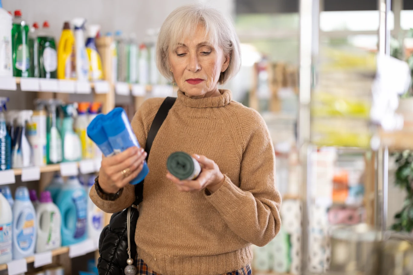 Starsza kobieta o siwych włosach w beżowym swetrze stoi w sklepie między regałami z chemikaliami gospodarskimi, trzymając dwa różne produkty i porównując ich etykiety.
