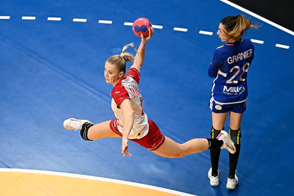 Zawodniczka w czerwonym stroju wykonuje dynamiczny skok z piłką ręczną w kierunku bramki na hali sportowej, w tle inna zawodniczka w niebieskim stroju.