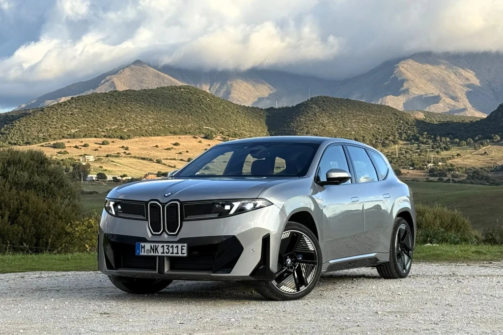BMW iX3 przejedzie na jednym ładowaniu więcej niż obiecuje producent. Zużycie nawet blisko 16 kWh/100 km. Nowoczesny SUV marki BMW w kolorze srebrnym stoi na żwirowej drodze z widokiem na zielone pagórki i góry w tle, częściowo skryte pod chmurami.