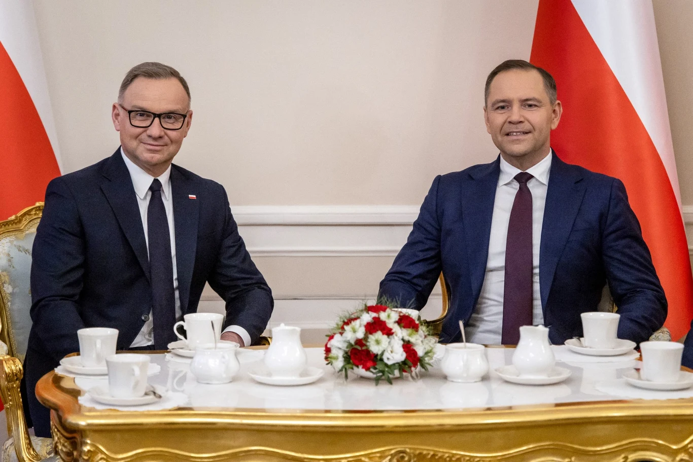 Dwóch elegancko ubranych mężczyzn siedzących przy ozdobnym stole w towarzystwie białych filiżanek, dzbanka i bukietu biało-czerwonych kwiatów, w tle biało-czerwona flaga.