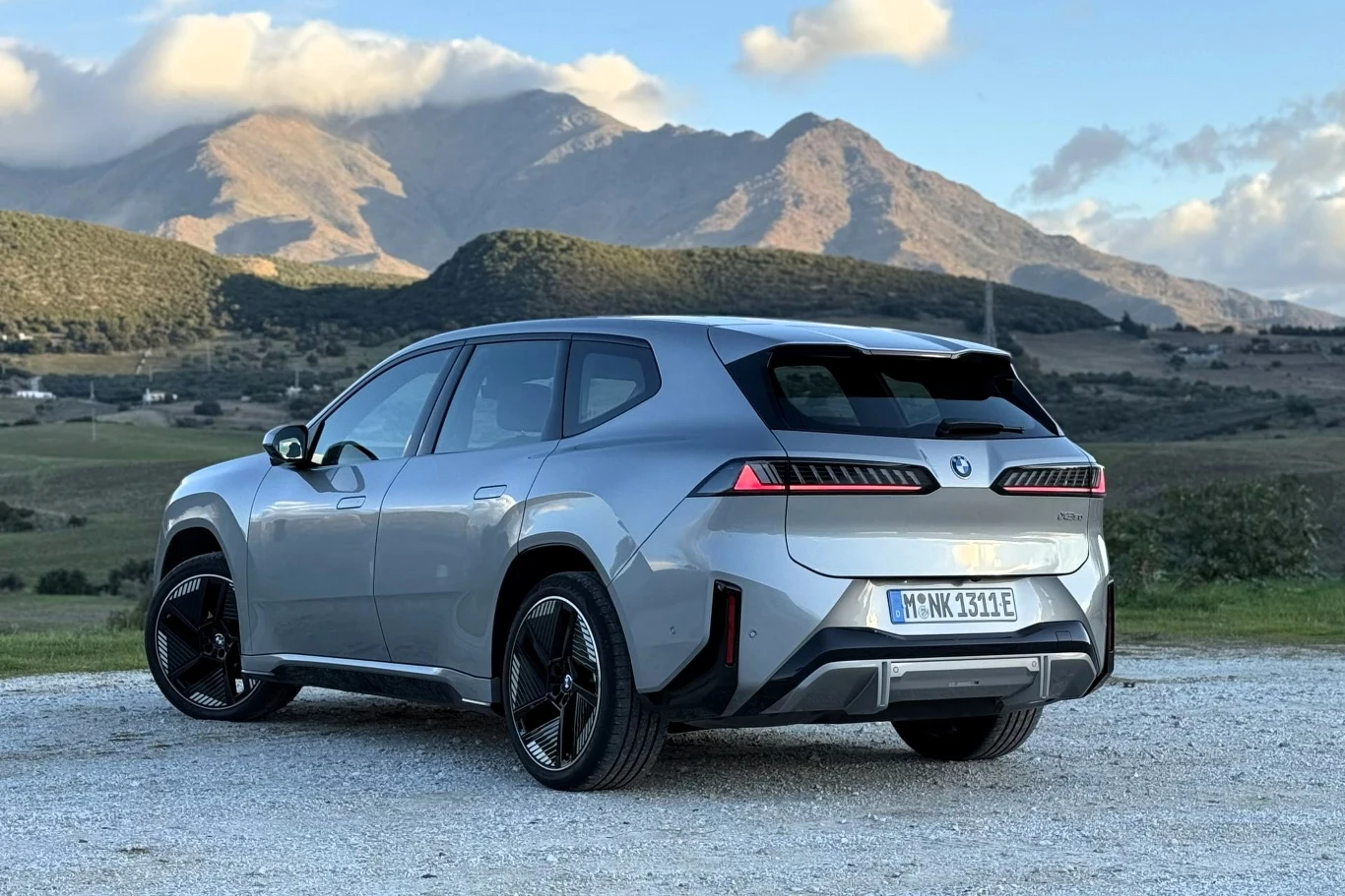 Nowoczesny srebrny SUV marki BMW zaparkowany na żwirowej nawierzchni, w tle rozległy krajobraz górski z wyraźnymi szczytami i chmurami na niebie.