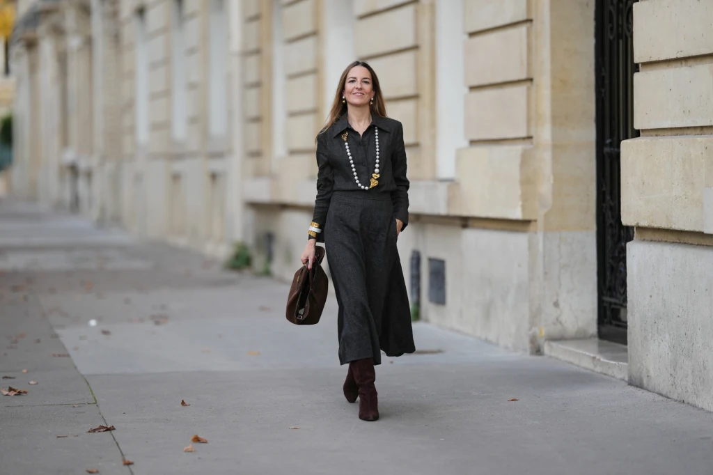 Kobieta ubrana w elegancką, ciemną sukienkę z długimi rękawami i naszyjnikiem, idzie chodnikiem przy jasnym, klasycznym budynku. Trzyma torebkę w dłoni i uśmiecha się lekko, wokół leżą pojedyncze liście.