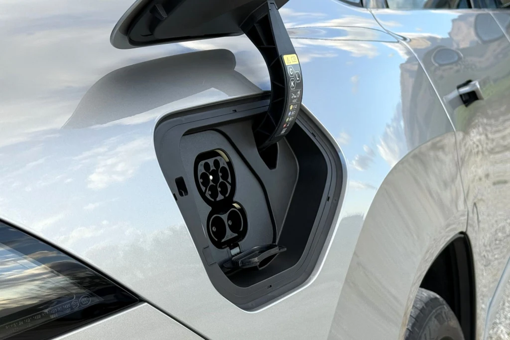 Akumulator BMW iX3 o pojemności 108,7 kWh pozwala na przejechanie do 805 km (zgodnie z danymi producenta) i obsługuje ładowanie prądem stałym o mocy 400 kW. Detal portu ładowania samochodu elektrycznego umieszczonego po lewej stronie srebrnego pojazdu z uchyloną klapką, na zewnątrz widoczne jasne, pochmurne niebo.