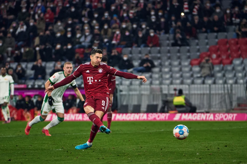 Robert Lewandowski za czasów gry w Bayernie Monachium Piłkarz w bordowym stroju wykonuje rzut karny podczas meczu piłki nożnej, w tle widać innych zawodników oraz trybuny wypełnione kibicami.