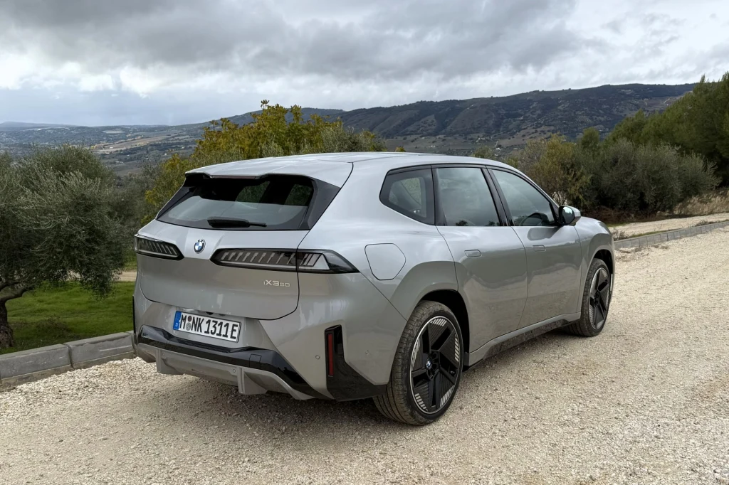 BMW deklaruje, że w czasie codziennej jazdy iX3 aż 98 proc. manewrów hamowania odbywa się za pomocą samej rekuperacji. Srebrny samochód marki BMW, model iX, zaparkowany na szutrowej drodze w otoczeniu zielonych drzew i wzniesień, widocznych w tle. Auto zwrócone tyłem, podkreślone nowoczesne linie nadwozia i designerskie felgi.