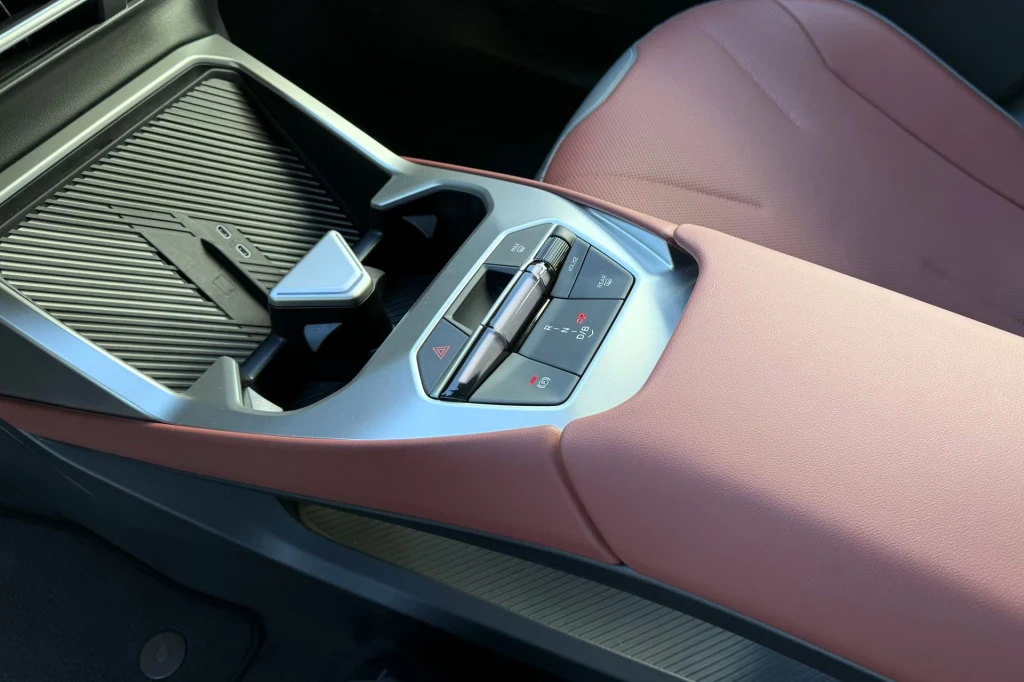 Wewnątrz BMW iX3 zostało kilka fizycznych przełączników, choćby dźwignia zmiany kierunku jazdy. Konsola środkowa nowoczesnego samochodu z różową tapicerką, panel sterowania przyciskami oraz pokrętłem, przestrzeń na drobne przedmioty i eleganckie wykończenie materiałów.