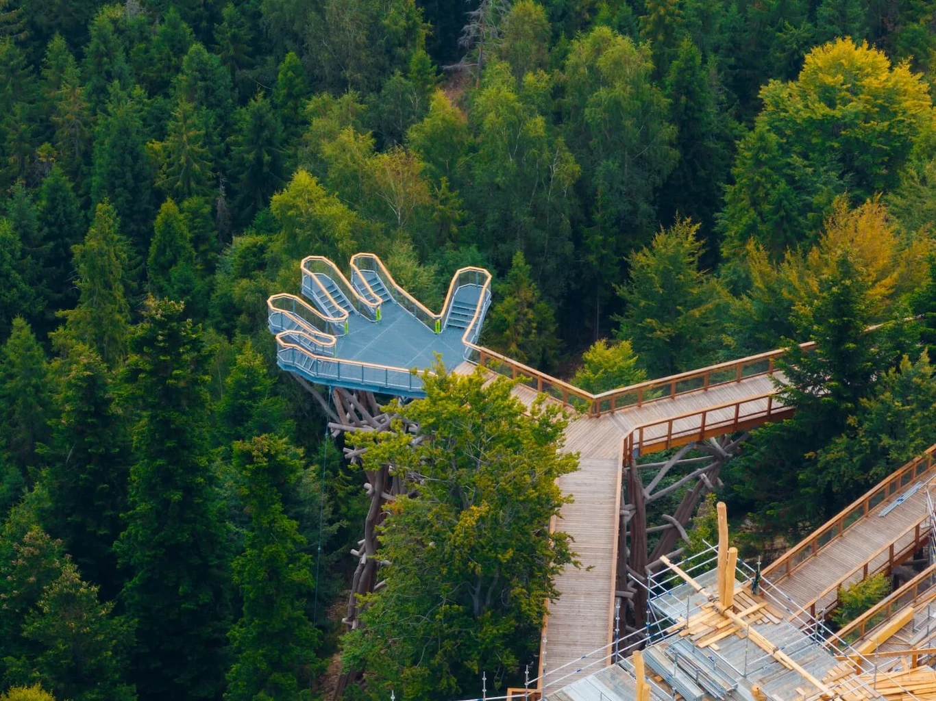Kładka prowadzi nad koronami gęstego lasu do platformy widokowej w kształcie ogromnej dłoni, otoczonej zielonymi drzewami.