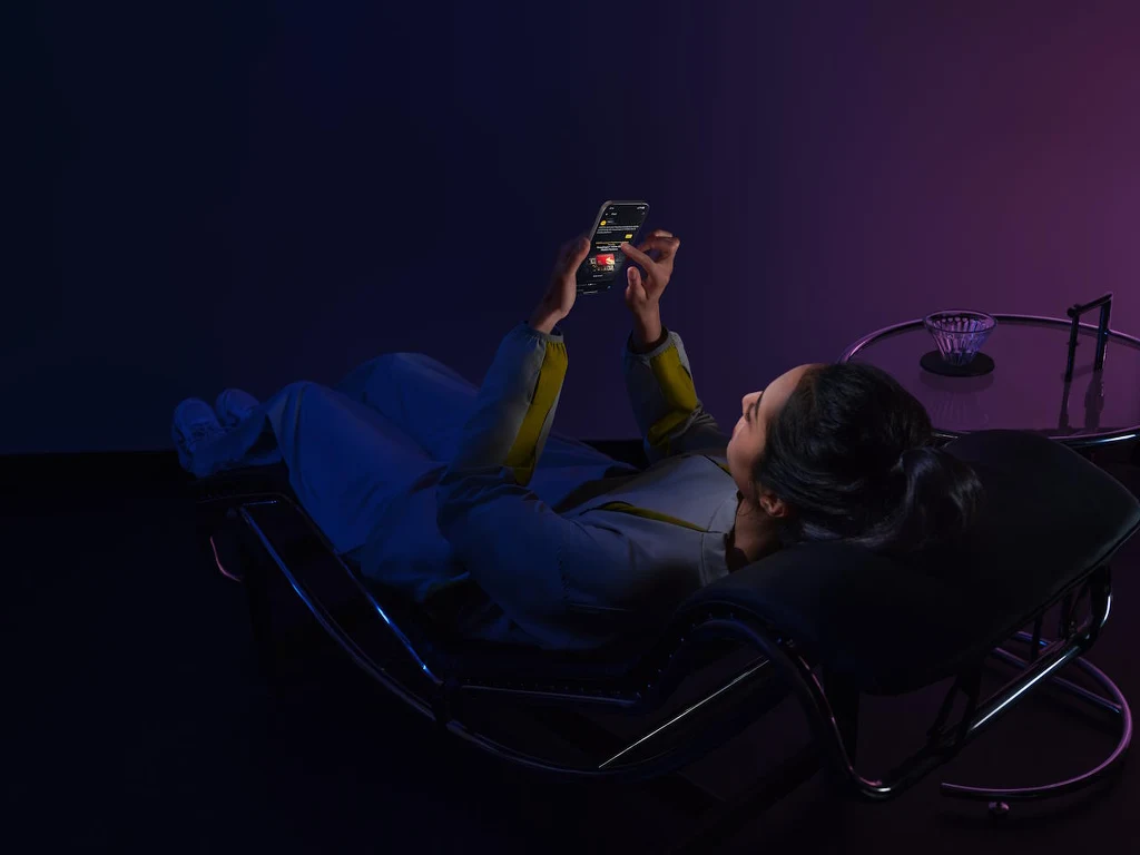 Mobilny gaming to duże obciążenie dla akumulatora, ale seria POCO F8 jest na to przygotowana Kobieta ubrana w casualowe ubrania leży na designerszym fotelu relaksacyjnym, trzyma w rękach otwarty smartfon z widocznym ekranem, obok znajduje się szklany stolik z popielniczką, całość oświetlona fioletowym światłem.