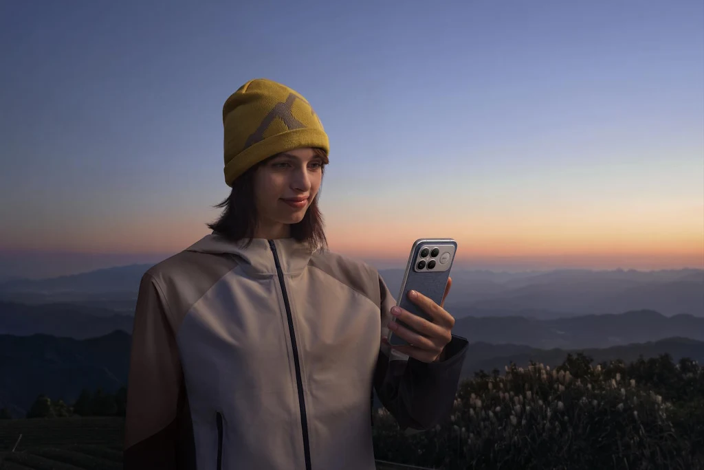 Seria POCO F8 to propozycja dla wymagających użytkowników Młoda kobieta w żółtej czapce i sportowej kurtce trzyma smartfona, stojąc na tle gór o zachodzie słońca, co podkreśla spokojną atmosferę i kontakt z technologią w otoczeniu natury.