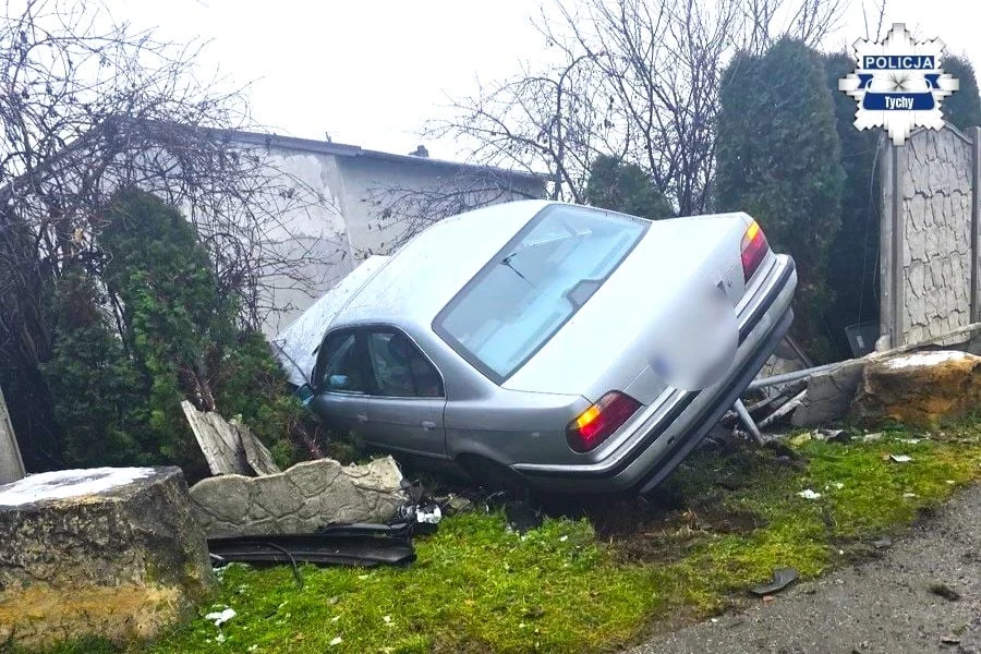 Srebrny samochód osobowy wjechał w betonowy płot i żywopłot, częściowo uniesiony, z uszkodzoną karoserią oraz elementami ogrodzenia wokół. W tle widoczna jest zabudowa gospodarcza i oznaczenie policji.