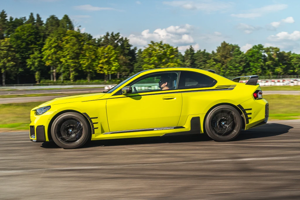 Jaskrawożółty sportowy samochód coupe jadący po torze asfaltowym, otoczony zielonymi drzewami i błękitnym niebem z lekkimi chmurami.