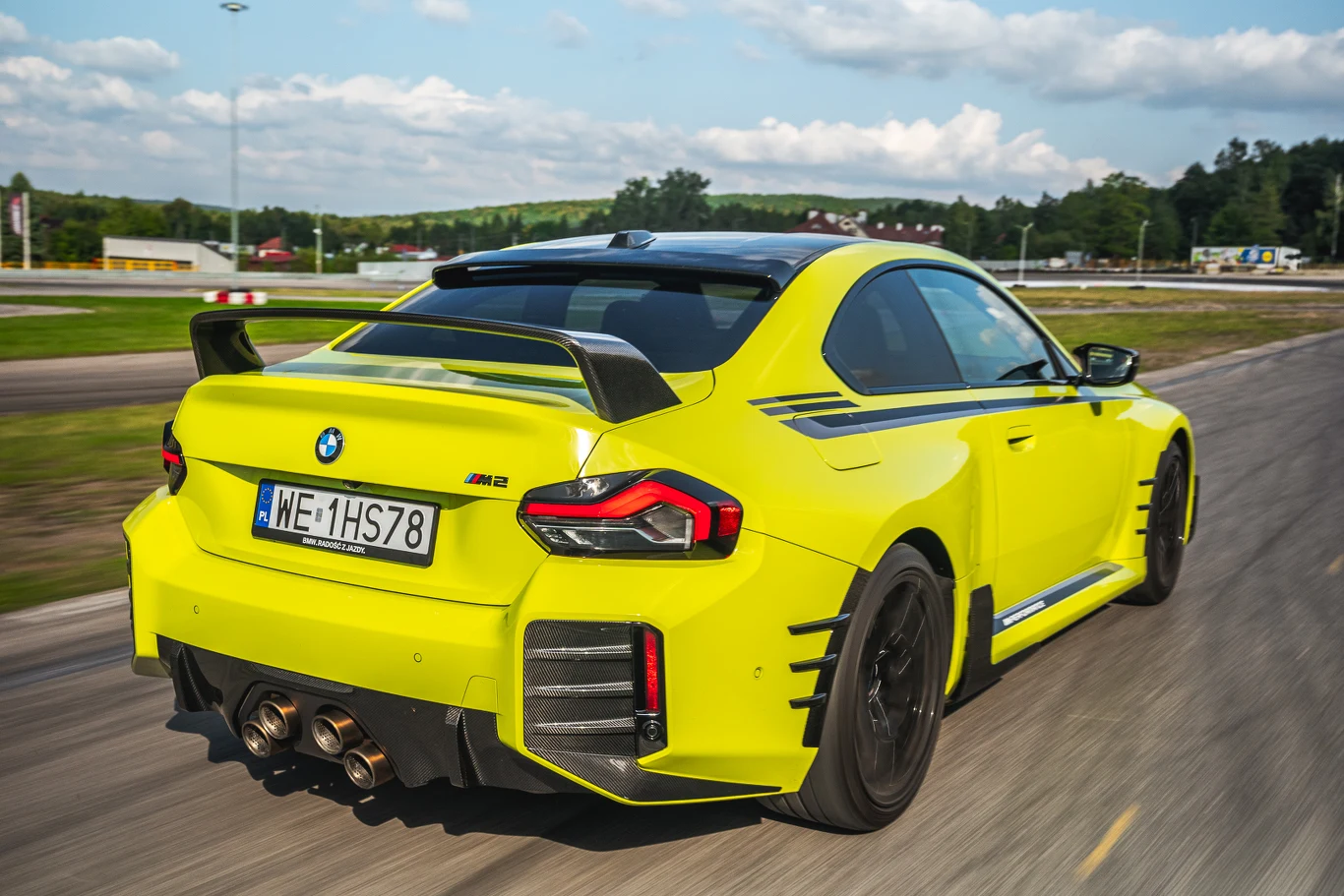 Niemal każdy czarny element widoczny na tym zdjęciu wykonany jest z karbonu - łącznie z dachem Jaskrawożółty samochód sportowy BMW M4 z aerodynamicznym spojlerem, czarnymi akcentami oraz sportowym wydechem porusza się po asfaltowym torze, wokół otoczony zielenią i niską zabudową.