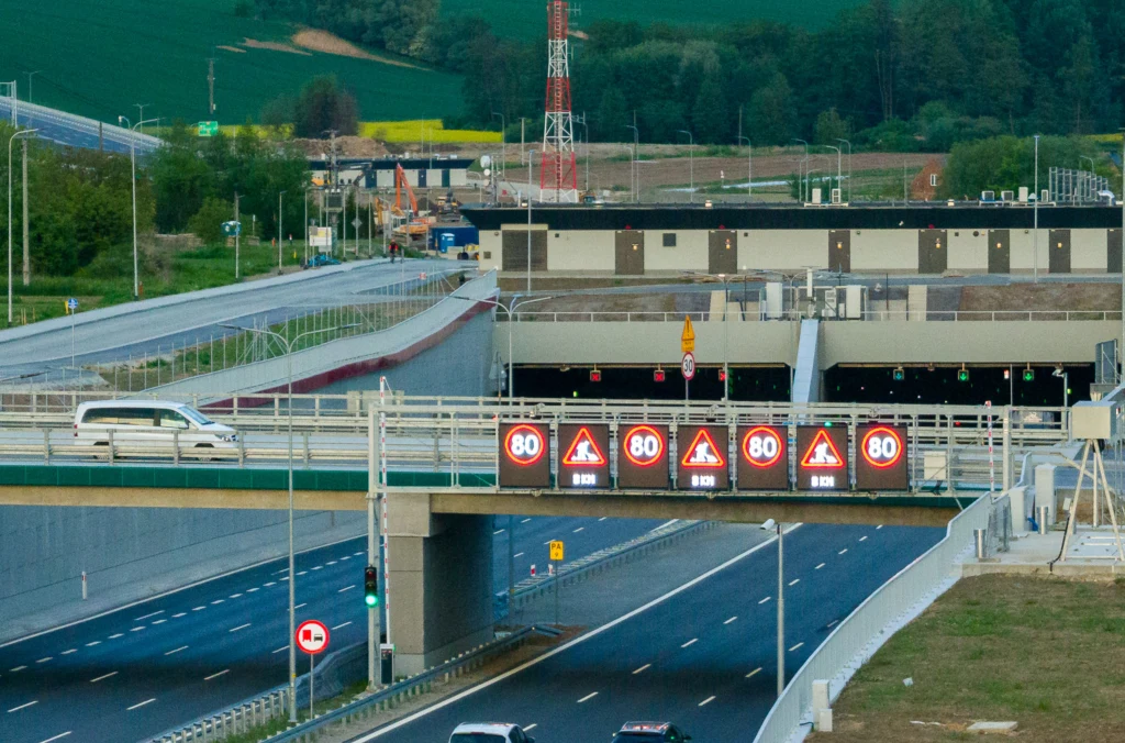 Wjazd do tunelu drogowego z widocznymi znakami ograniczenia prędkości do 80 km/h oraz oznaczeniami ostrzegawczymi na elektronicznych tablicach. Most nad wjazdem, samochody poruszające się po obu drogach i otoczenie z zielonymi polami w tle.