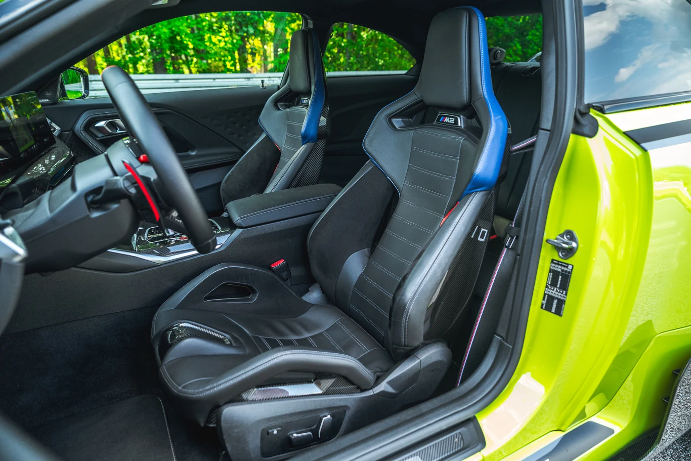 Kubełkowe fotele M Carbon wymagają dopłaty aż 22 400 zł, ale prezentują się doskonale i takie też zapewniają wrażenia podczas jazdy Wnętrze nowoczesnego samochodu sportowego z dwoma fotelami kubełkowymi, wykończonymi skórą i materiałem, sportowa kierownica oraz elementy wykończenia w kolorze kontrastującym z tapicerką. Za oknami widoczny las i intensywne światło dzienne.