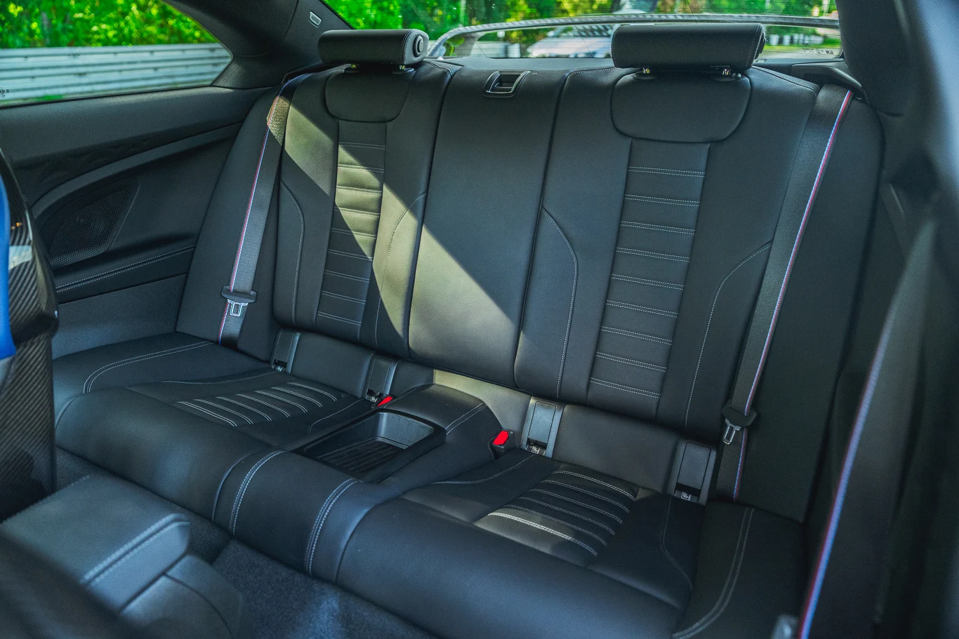 Tylna kanapa w BMW M2 nie wygląda źle, ale miejsca jest tu bardzo mało. Nie ma tu podłokietnika, a jedynie otwór na narty Skórzana, czarna tylna kanapa w nowoczesnym samochodzie, z wyraźnymi przeszyciami, regulowanymi zagłówkami oraz dwoma pasami bezpieczeństwa, widoczna sylwetka drzwi oraz elementy wnętrza auta.