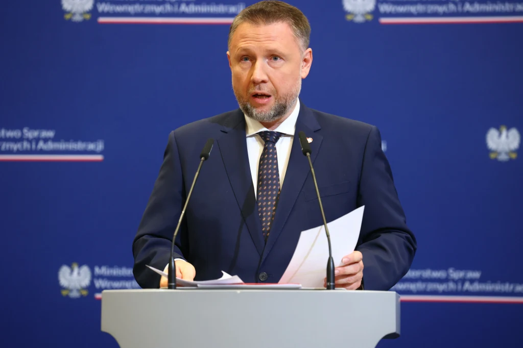 Mężczyzna w garniturze przemawia przy mównicy na tle niebieskiej planszy z napisem Ministerstwo Spraw Wewnętrznych i Administracji oraz polskimi godłami po obu stronach.