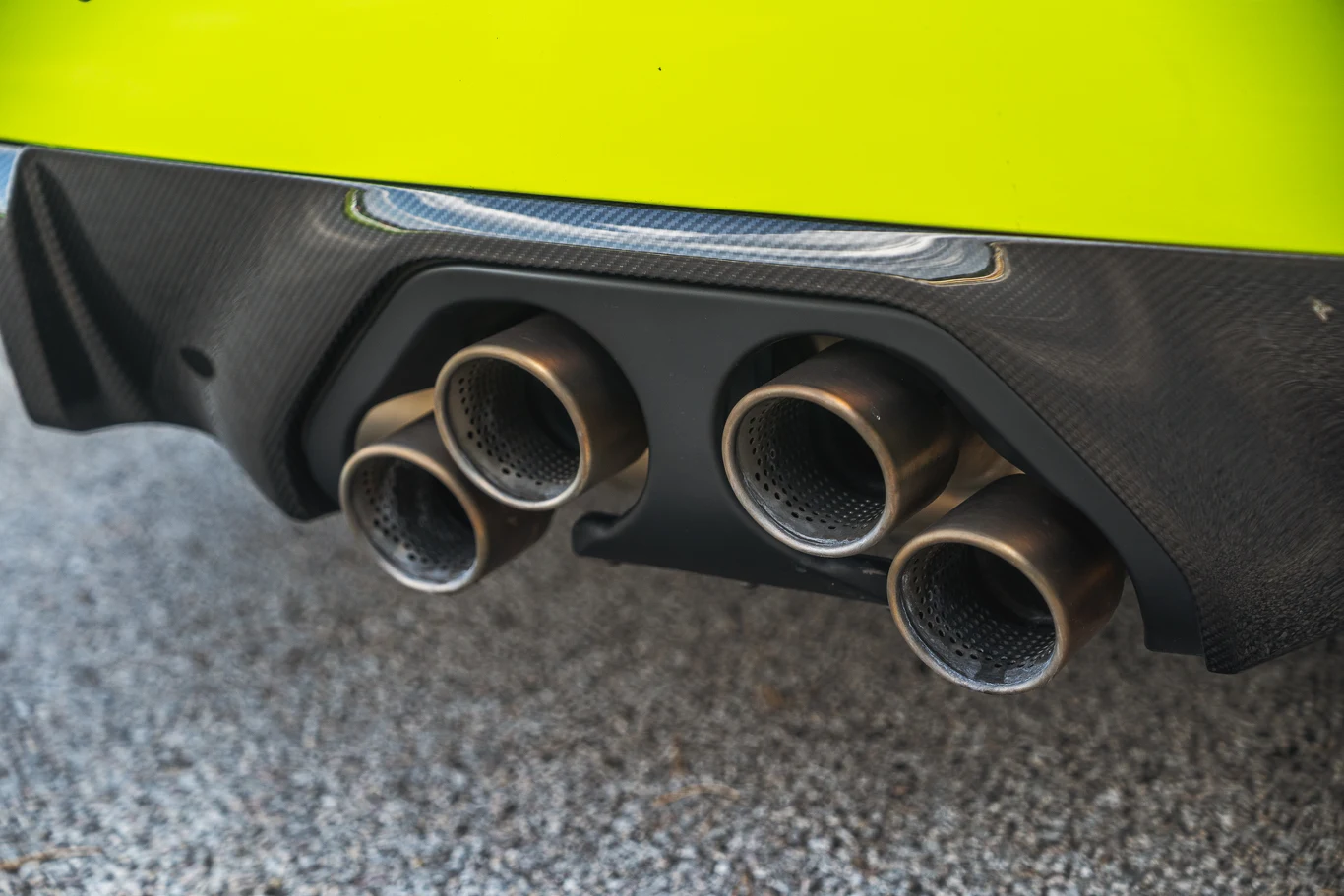 Akcesoryjny wydech z centralnymi końcówkami wygląda jakby pluł ogniem przy każdym dodaniu gazu. Niestety nie robi tego, ale brzmi bardzo rasowo, szczególnie jak na dzisiejsze standardy Cztery metalowe rury wydechowe zamontowane w dolnej części sportowego samochodu w jaskrawożółtym kolorze, na tle asfaltowej nawierzchni.