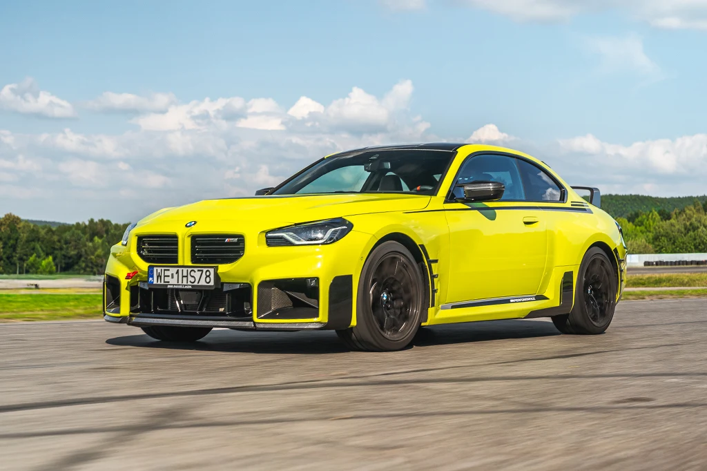 Żółte sportowe coupe marki BMW jadące po torze wyścigowym, na tle zielonej roślinności oraz nieba z rozproszonymi chmurami.