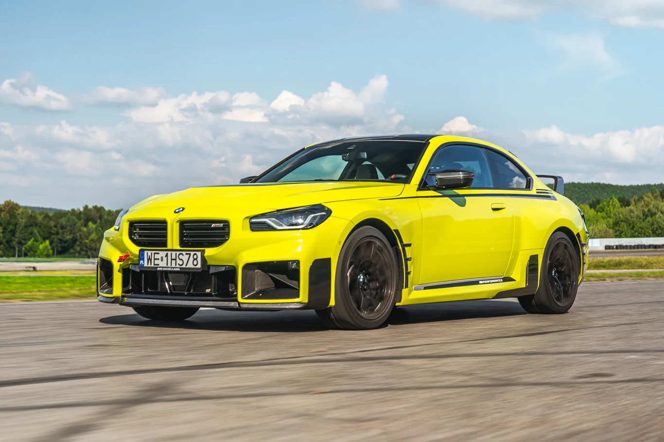 BMW M2 wzbogacone o dodatki M Performance nabiera znacznie ostrzejszego charakteru Żółte sportowe coupe marki BMW jadące po torze wyścigowym, na tle zielonej roślinności oraz nieba z rozproszonymi chmurami.