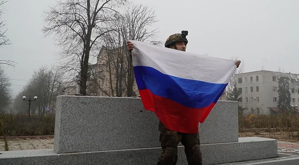 Żołnierz w mundurze wojskowym trzyma dużą flagę Rosji, stojąc przed betonowym podestem w miejskim otoczeniu z drzewami i budynkami w tle, przy pochmurnej pogodzie.
