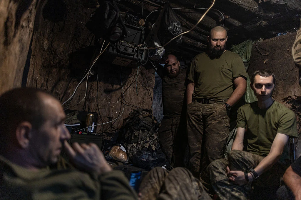 Grupa żołnierzy w wojskowych ubraniach siedzi i stoi w ziemiance, otoczeni sprzętem wojskowym, kablami i drewnianymi elementami konstrukcji, atmosfera skupienia i zmęczenia.