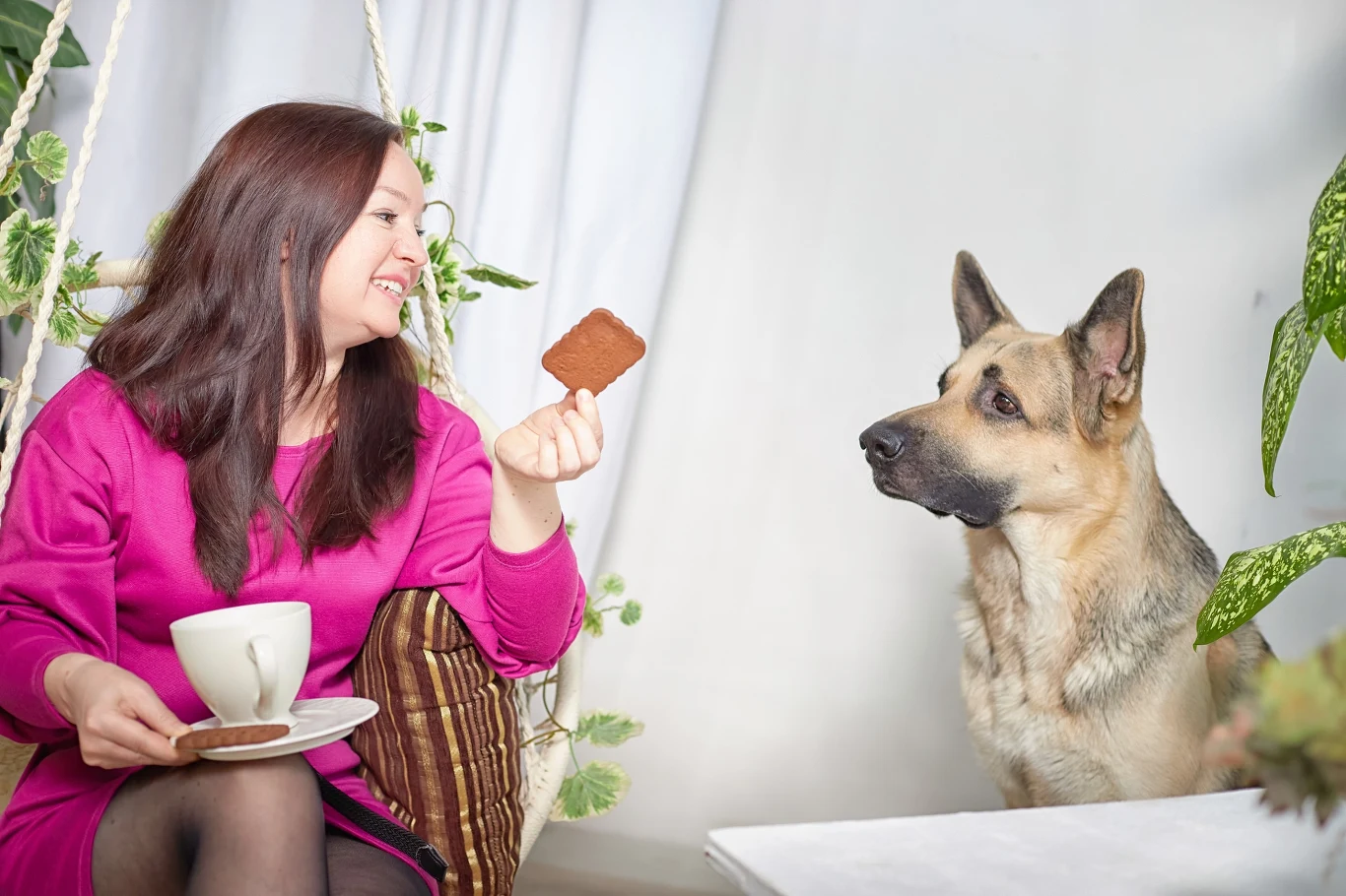 Kobieta w różowej sukience siedzi na huśtawce z filiżanką i ciastkiem w ręku. Przed nią siedzi owczarek niemiecki, patrząc uważnie na trzymane ciastko.