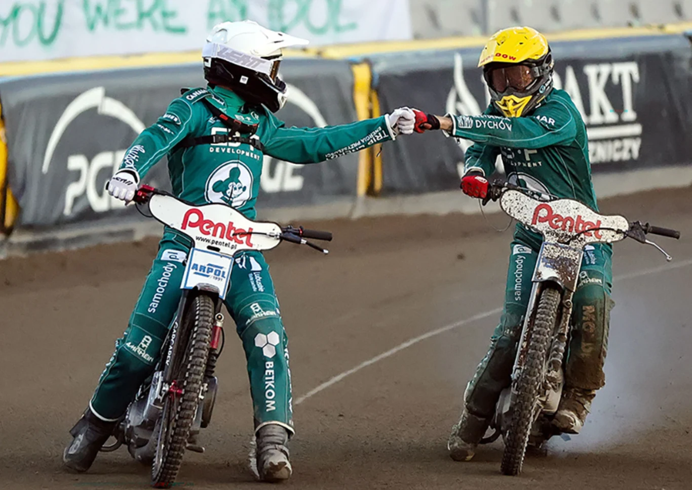 Dwóch żużlowców w zielonych kombinezonach i kaskach jedzie obok siebie po torze, wykonując gest przybicia piątki, podkreślający sportową współpracę i koleżeństwo.