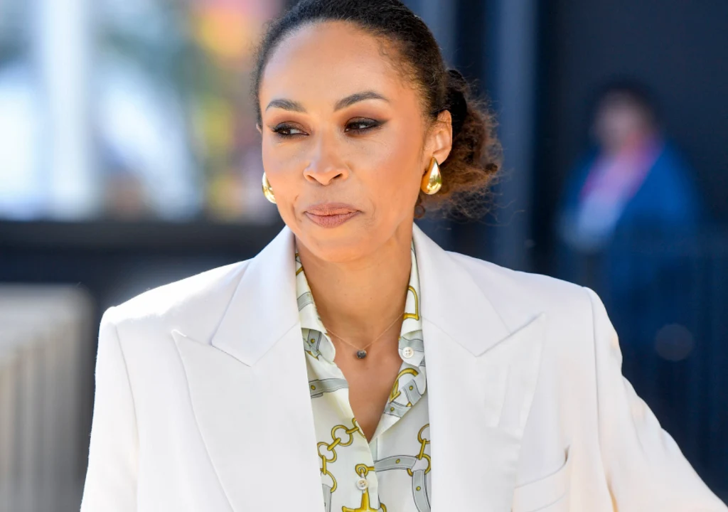 Kobieta o ciemnych włosach spiętych w kok ubrana w elegancki biały żakiet oraz koszulę z ozdobnym nadrukiem, nosząca złote kolczyki i delikatny naszyjnik, znajduje się na zewnątrz w jasnym świetle dziennym.