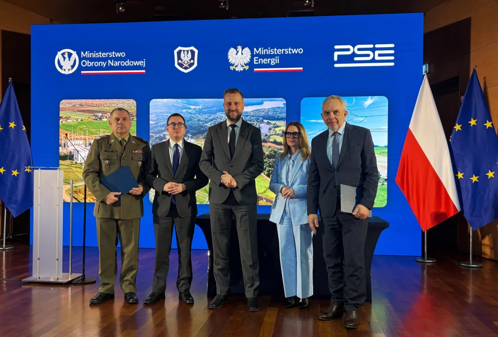 Grupa pięciu osób stojąca przed niebieskim tłem z logotypami Ministerstwa Obrony Narodowej, Ministerstwa Energii, PSE oraz flagami Unii Europejskiej i Polski, wszyscy ubrani formalnie, z dokumentami w dłoniach.
