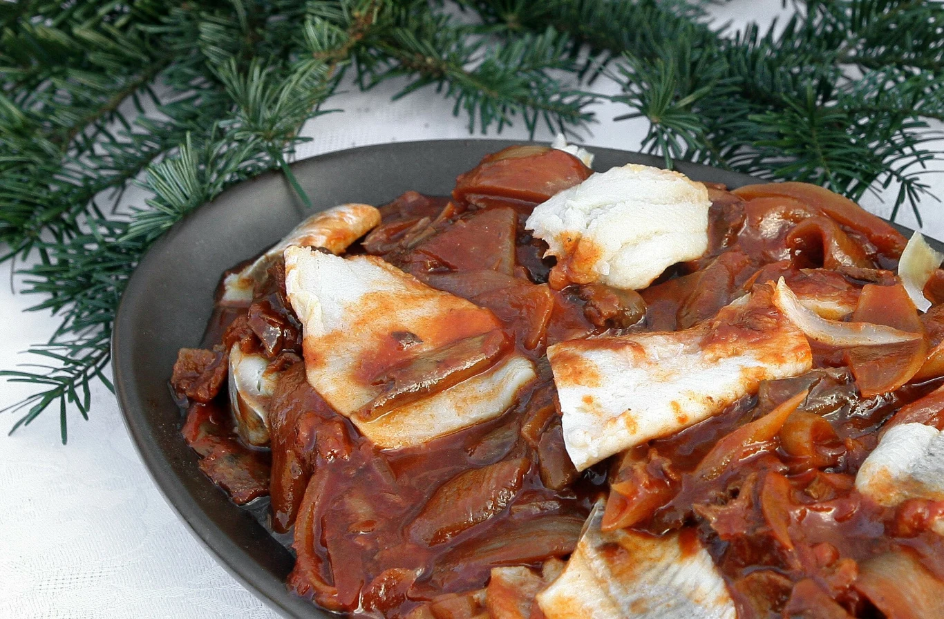 Śledzie po grecku to tradycyjne danie wigilijne, które warto zrobić nieco wcześniej Kawałki śledzia zanurzone w aromatycznym sosie pomidorowym z cebulą, podane na ciemnym talerzu. W tle widoczne zielone gałązki świerku, sugerujące świąteczny charakter potrawy.